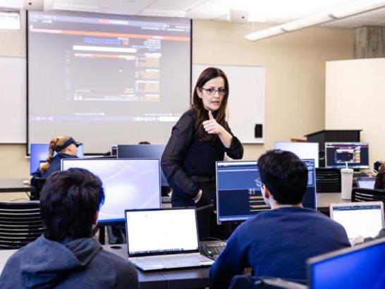 A professor gives students a thumbs up as she walks through a row of desks in the classroom.